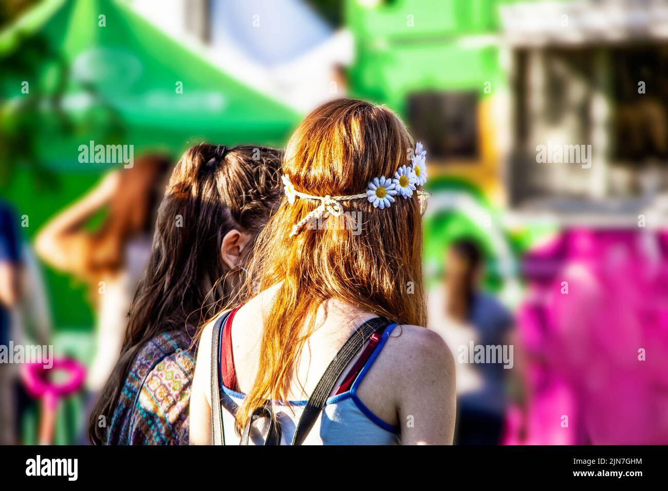 Two girls - one with red hair and daisy headband - wait in line at food truck at carnival - bright colors of pink and green Stock Photo