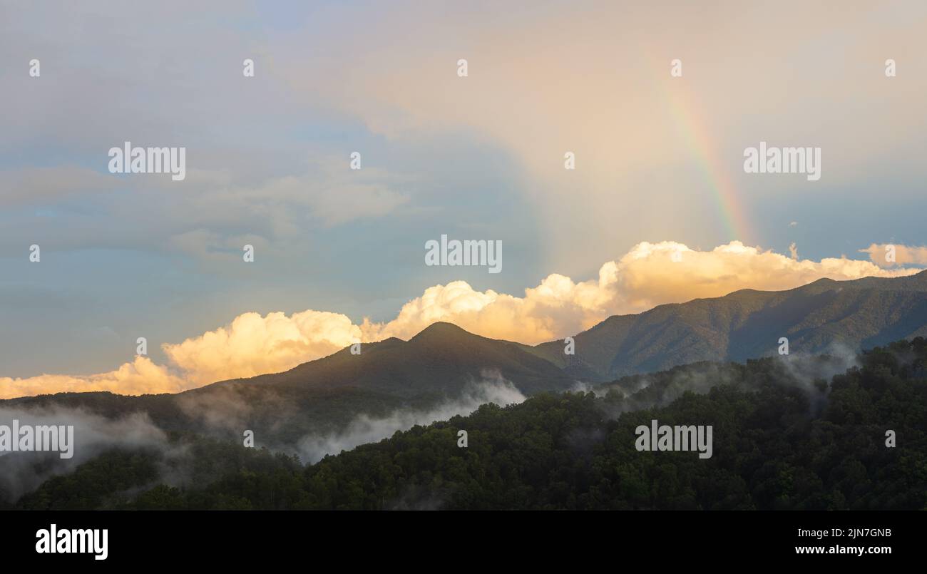 A sunset with a rainbow and large puffy clouds in the Great Smoky Mountain National Park. Smoke lingers in the mountains after a heavy storm. Stock Photo