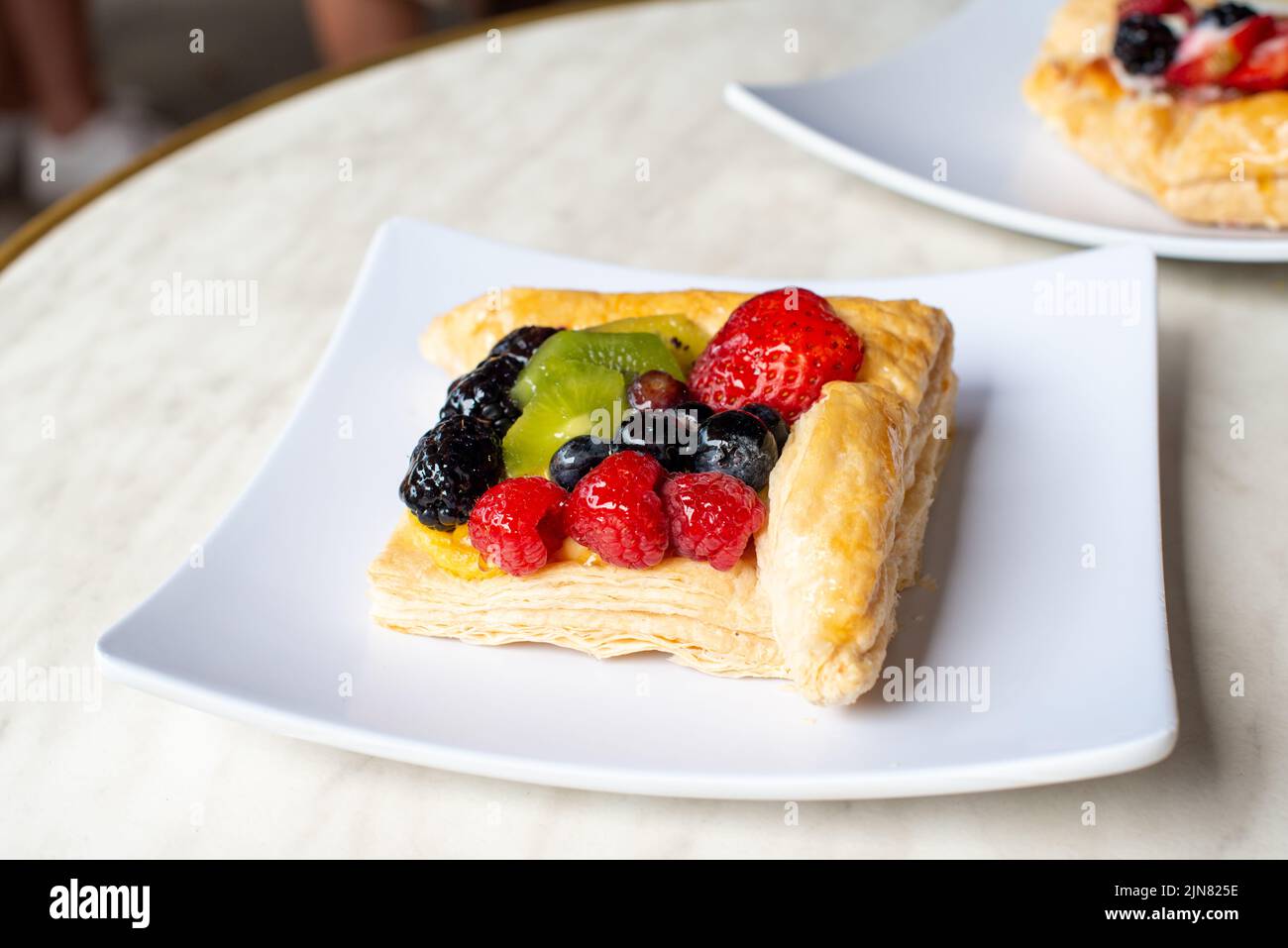 A square fruit filled French puff pastry on a white square plate. The cream and custard filled dessert has strawberries, kiwi, and blackberries on top Stock Photo