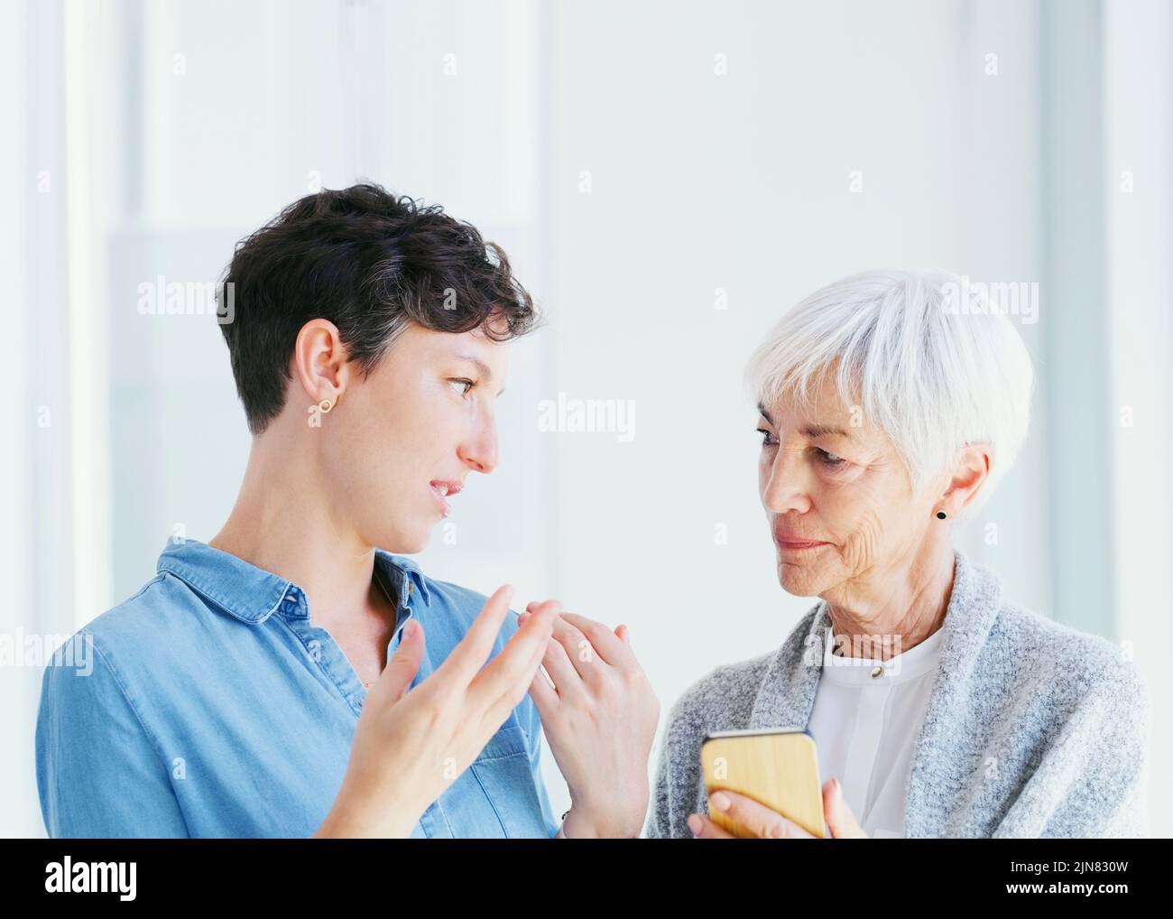 This is how social media works. a senior woman holding a cellphone while talking with her attractive young daughter while at home. Stock Photo