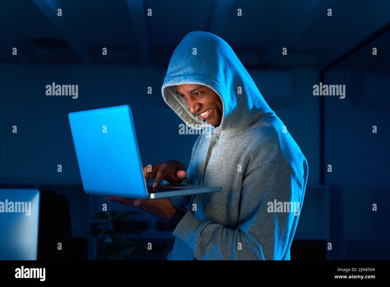 Firewall breached. a young male hacker smiling while using a laptop in the dark. Stock Photo