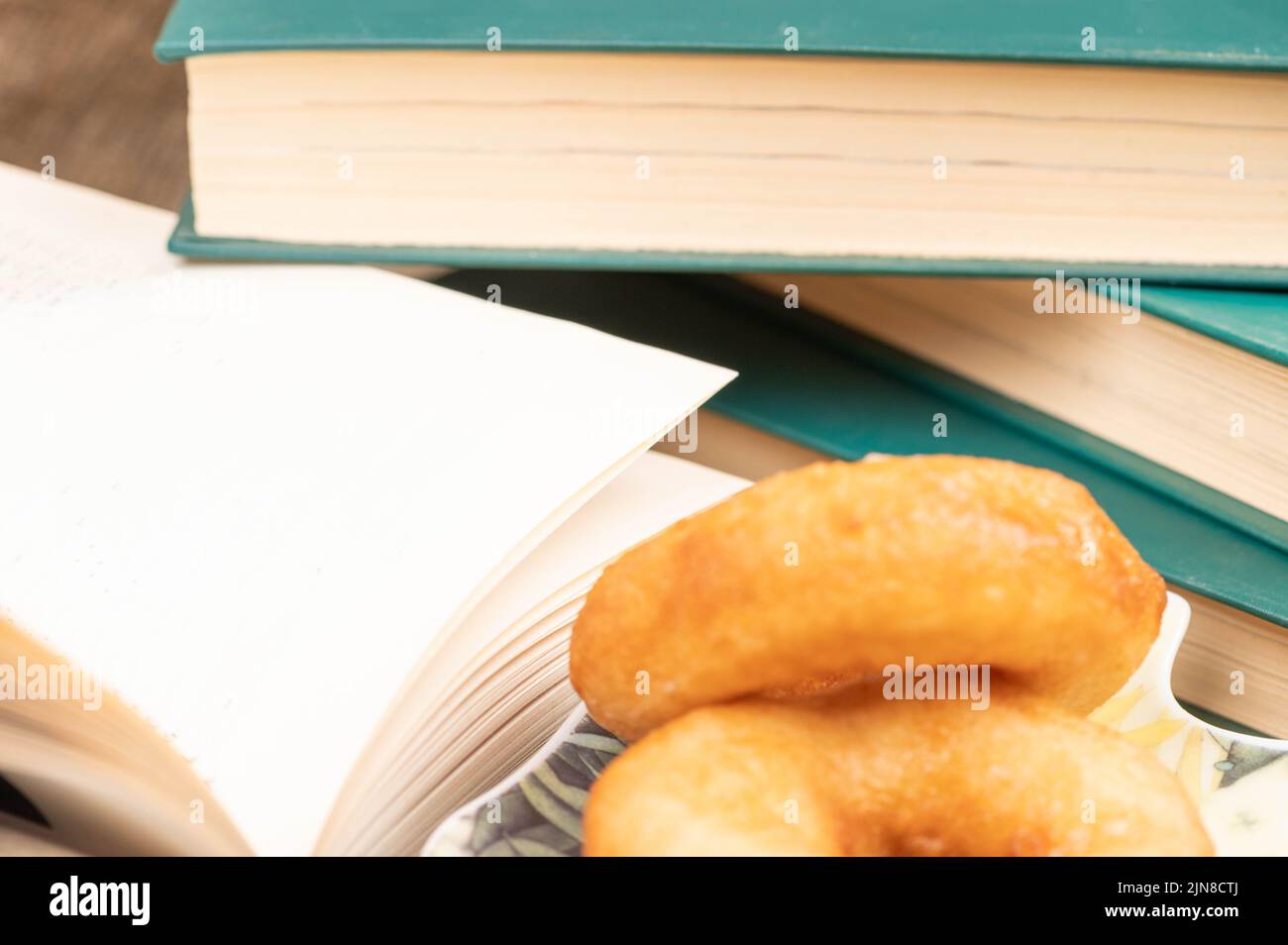 A cup of strong tea on a saucer, donuts on a plate and a stack of books on the table. Stock Photo