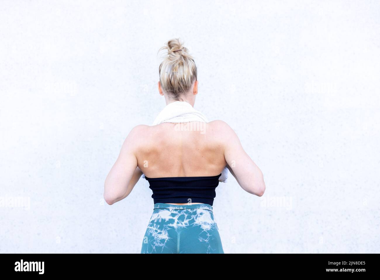Half Body Rear View Shot of a Gym Fit Caucasian sporty woman dressed in sportswear and with her hair up holding a towl around her neck with her hands and standing in the gym. In background wall, backs turned. High quality photo Stock Photo