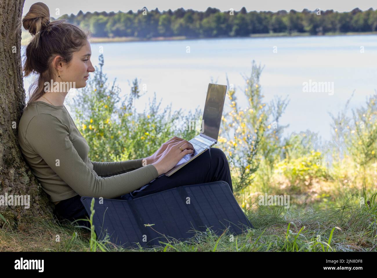 Work from anywhere. Remote freelancer work in nature using renewable energy via a foldable solar panel. Young woman, female freelancer working with laptop with Beautiful view of forest and lake. freelancer paradise, freedom of teleworking. distance. High quality photo Stock Photo