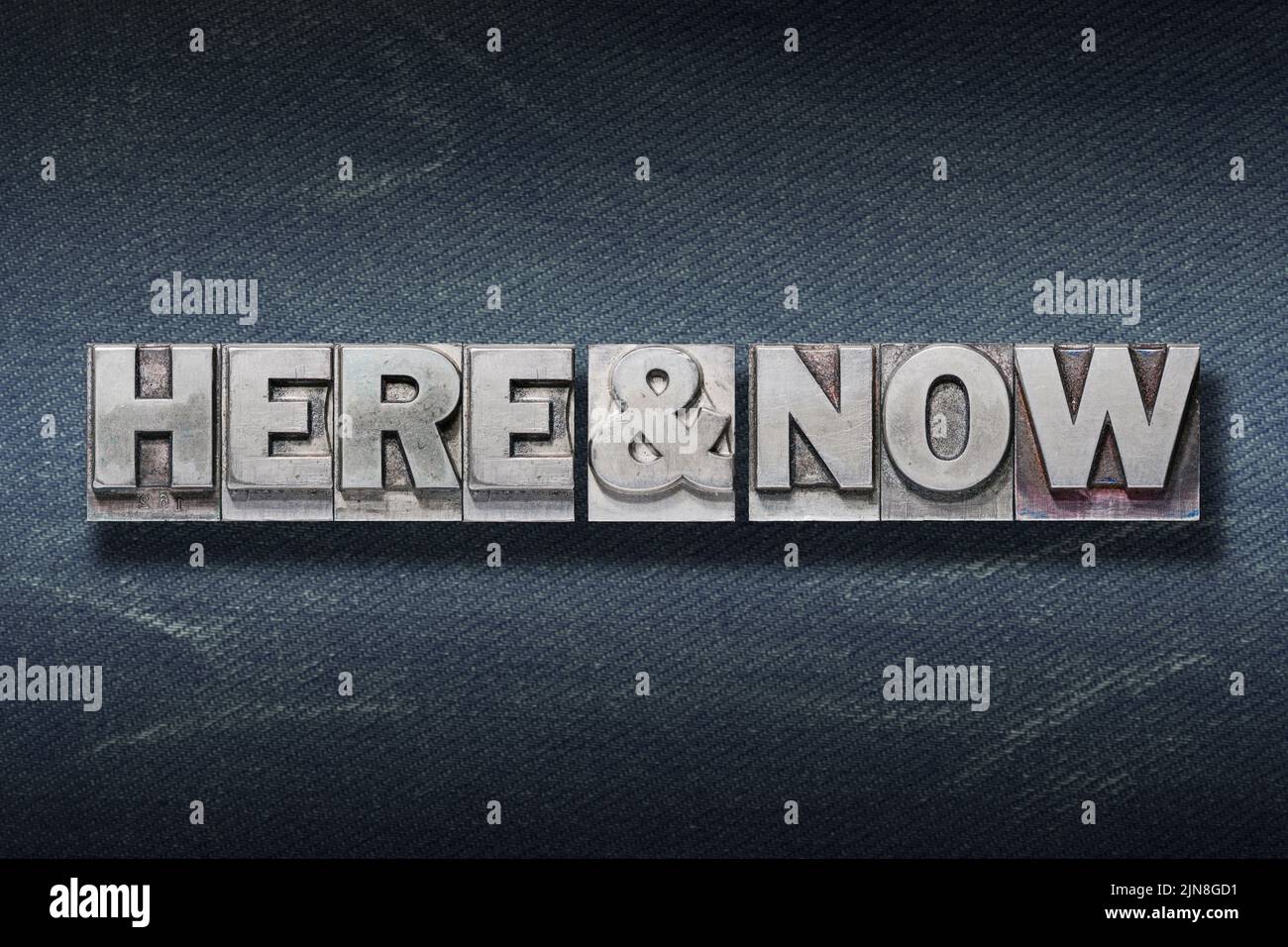 here and now phrase made from metallic letterpress on dark jeans background Stock Photo