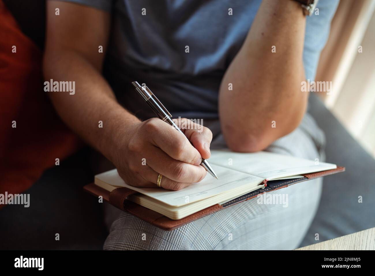 Photo closeup of adult man sitting on soft chair and writing notes by hand in notebook with pen. Business person resolving work issues. Remote job Stock Photo