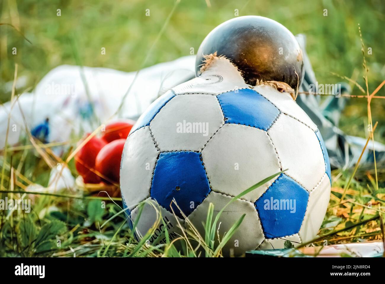 Burst soccer ball lies in pile of garbage on green grass in city. Torn ball thrown as rubbish after playing football game close view Stock Photo