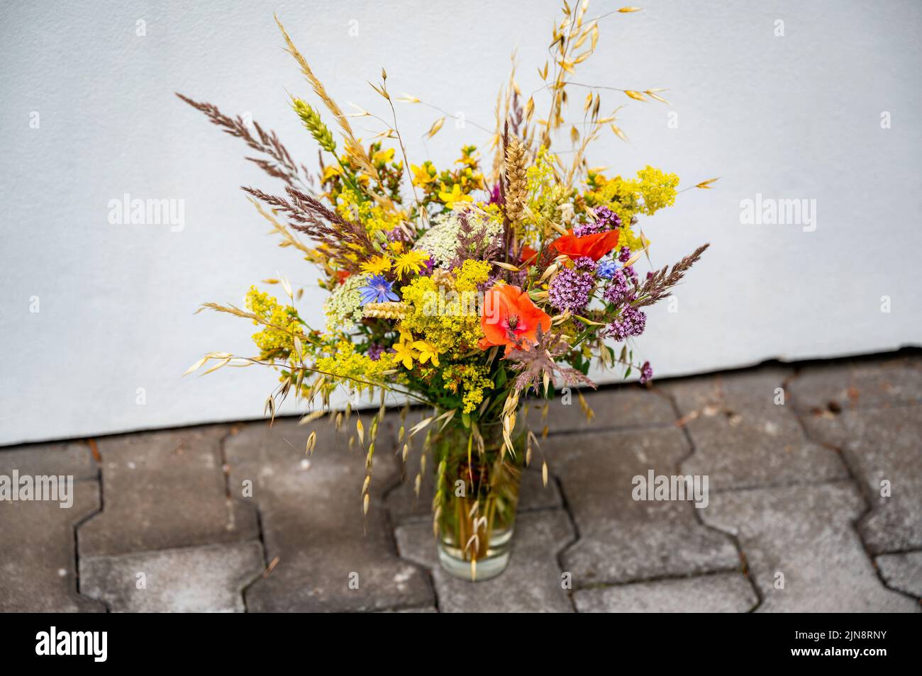 Bouquet of wild meadow and field flower in glass on paving and white backgroud wall. Stock Photo