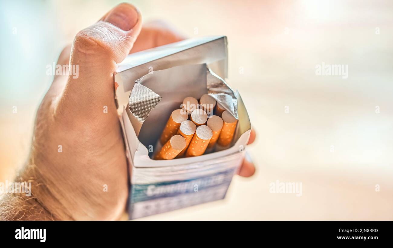 Hand of man holding pack of cigarettes on blurred background. Guy opens paper pack to smoke. Smoking addiction and bad habit close view Stock Photo
