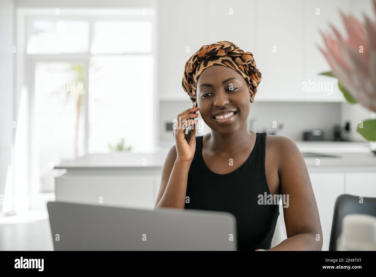 Beautiful young African woman wearing tradition headscarf. Sitting at home working on laptop. On phone call smiling Stock Photo