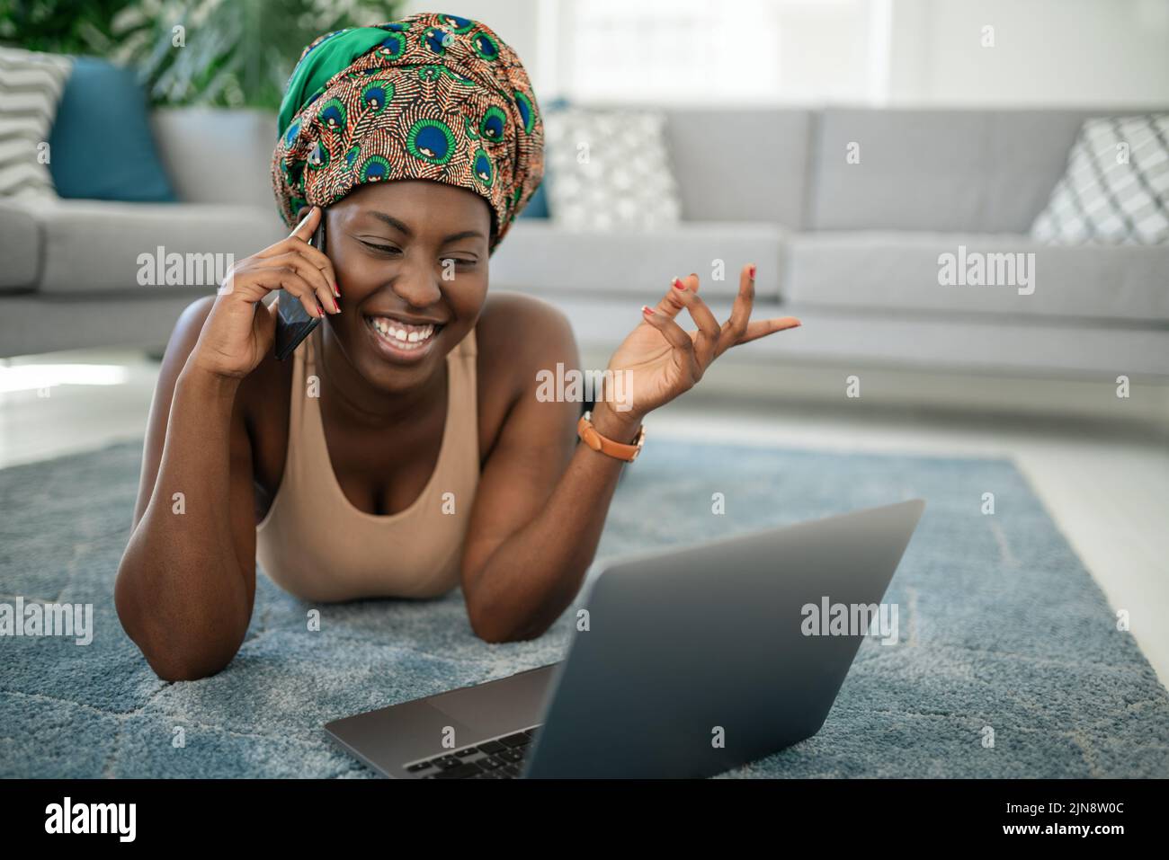 Beautiful African woman at home, smiling and laughing, using mobile smart phone, wearing traditional headscarf, surprised and happy Stock Photo