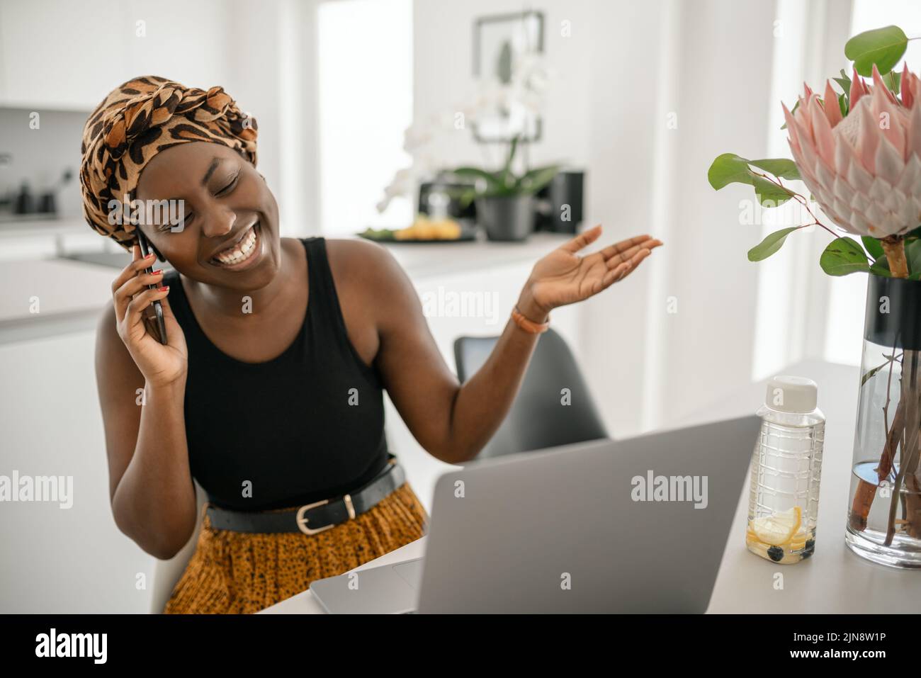 Beautiful young African woman wearing tradition headscarf. Sitting at home working on laptop. On phone call smiling and gesturing with hand Stock Photo