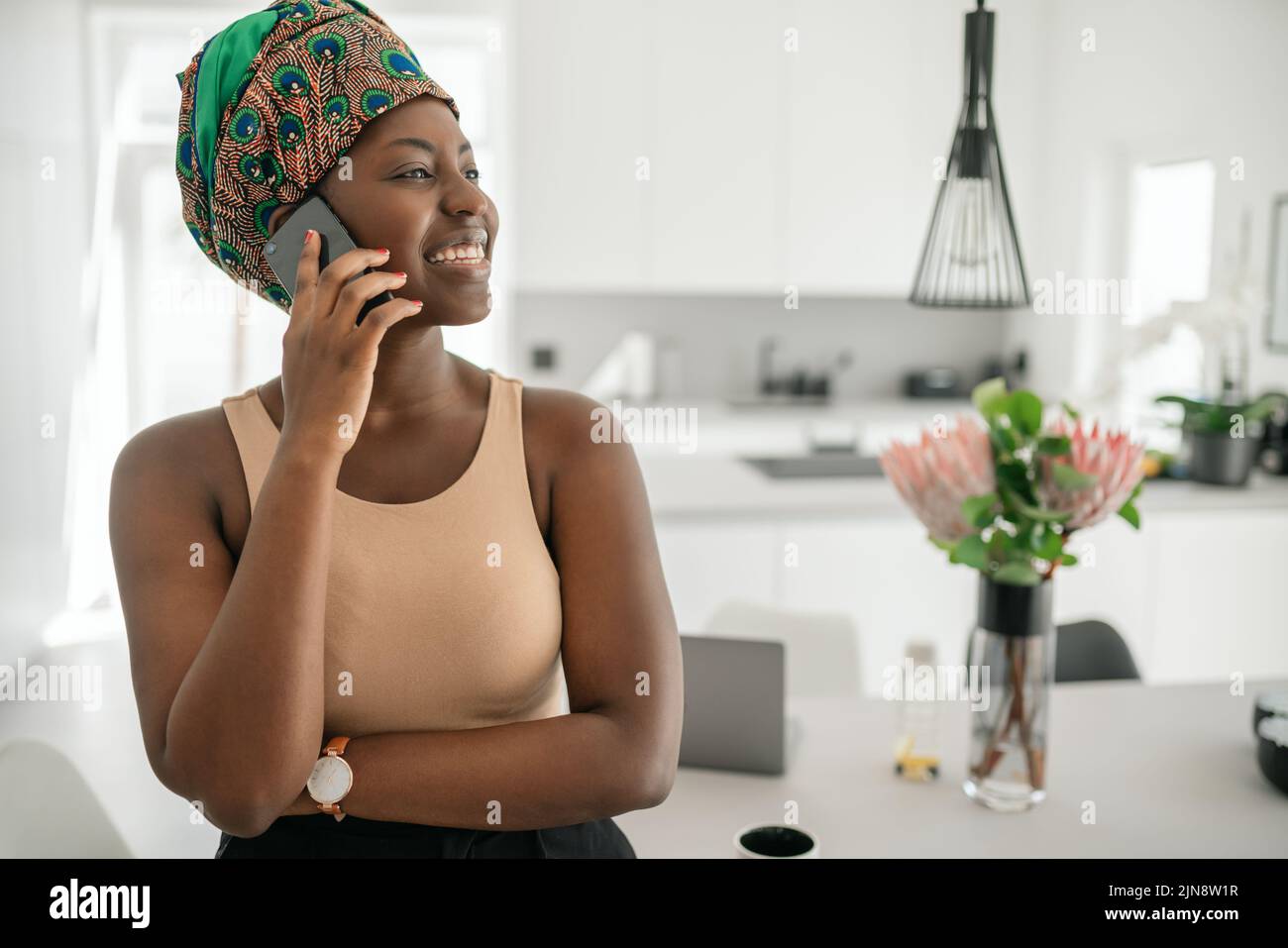 Beautiful African woman at home, smiling, using mobile smart phone, wearing traditional headscarf and looking into distance with copy space Stock Photo
