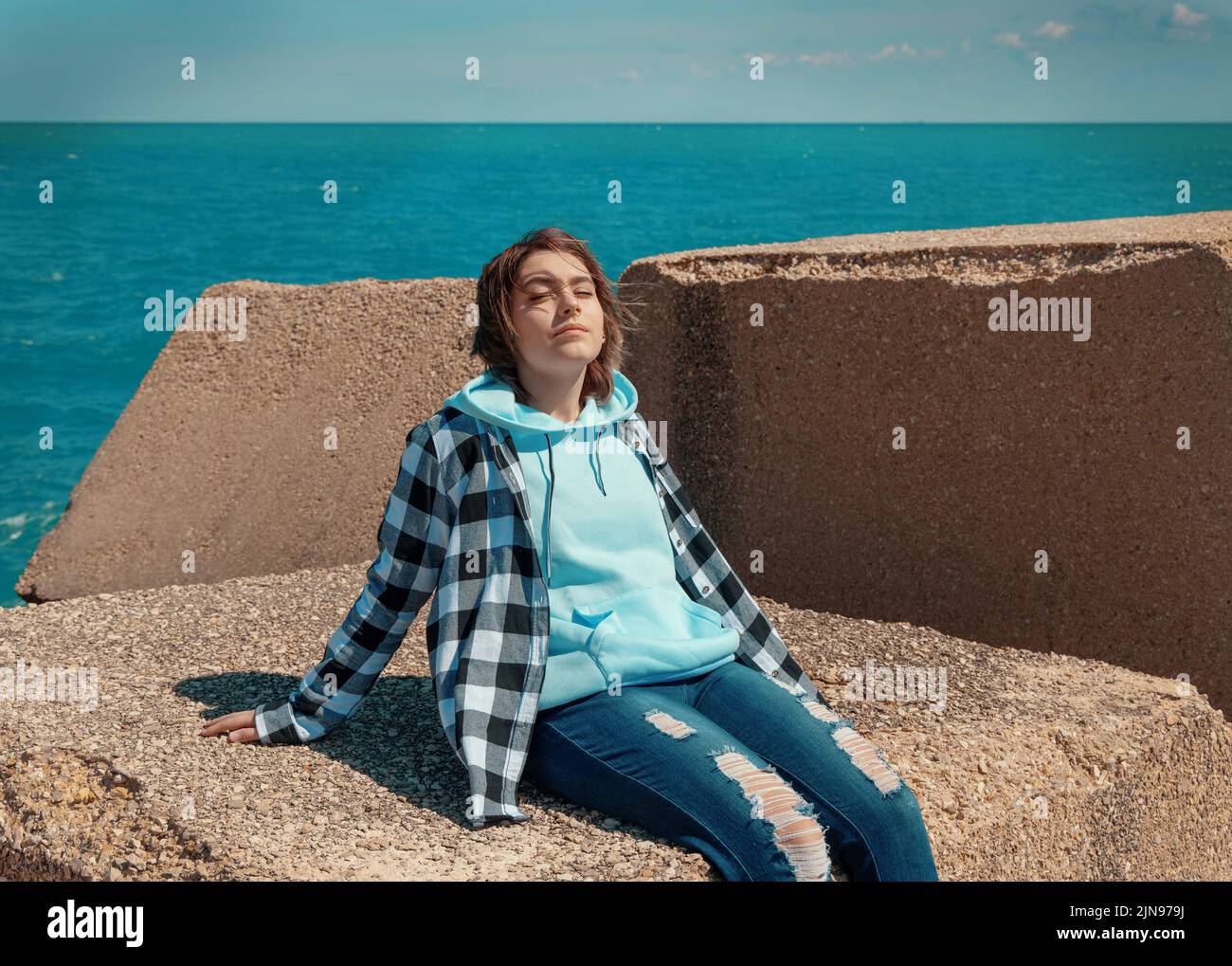Relaxed Teenager girl in light blue hoodie and plaid shirt. Smiling Teen girl sitting outdoor and enjoying the sunny day. Adolescence concept Stock Photo