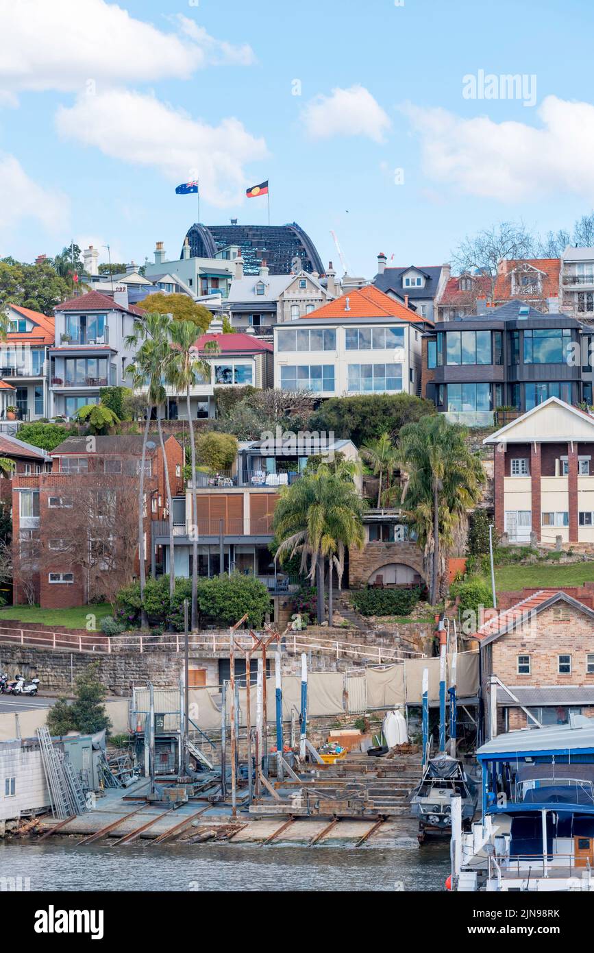 The flags and arch of the Sydney Harbour Bridge peak over the hill behind houses in Kirribilli and Sydney Harbour Slipways Pty Ltd on Careening Cove Stock Photo