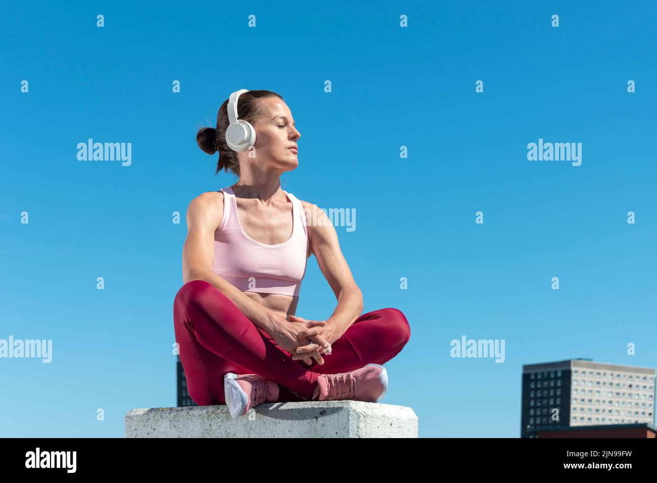 attractive fit woman sitting crossed legged slistening to music with headphones outdoors in the sun, urban setting. Stock Photo