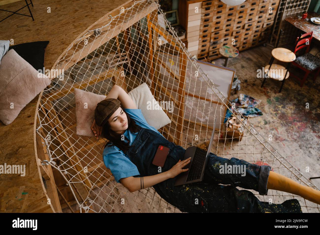 Young woman artist using laptop while resting in hammock at her studio indoors Stock Photo