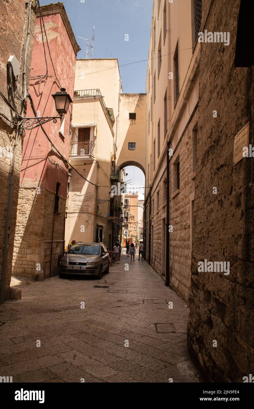 Old town Bari, narrow alleyway with arch and kids playing in the distance, parked car, strong sun and shadows Stock Photo