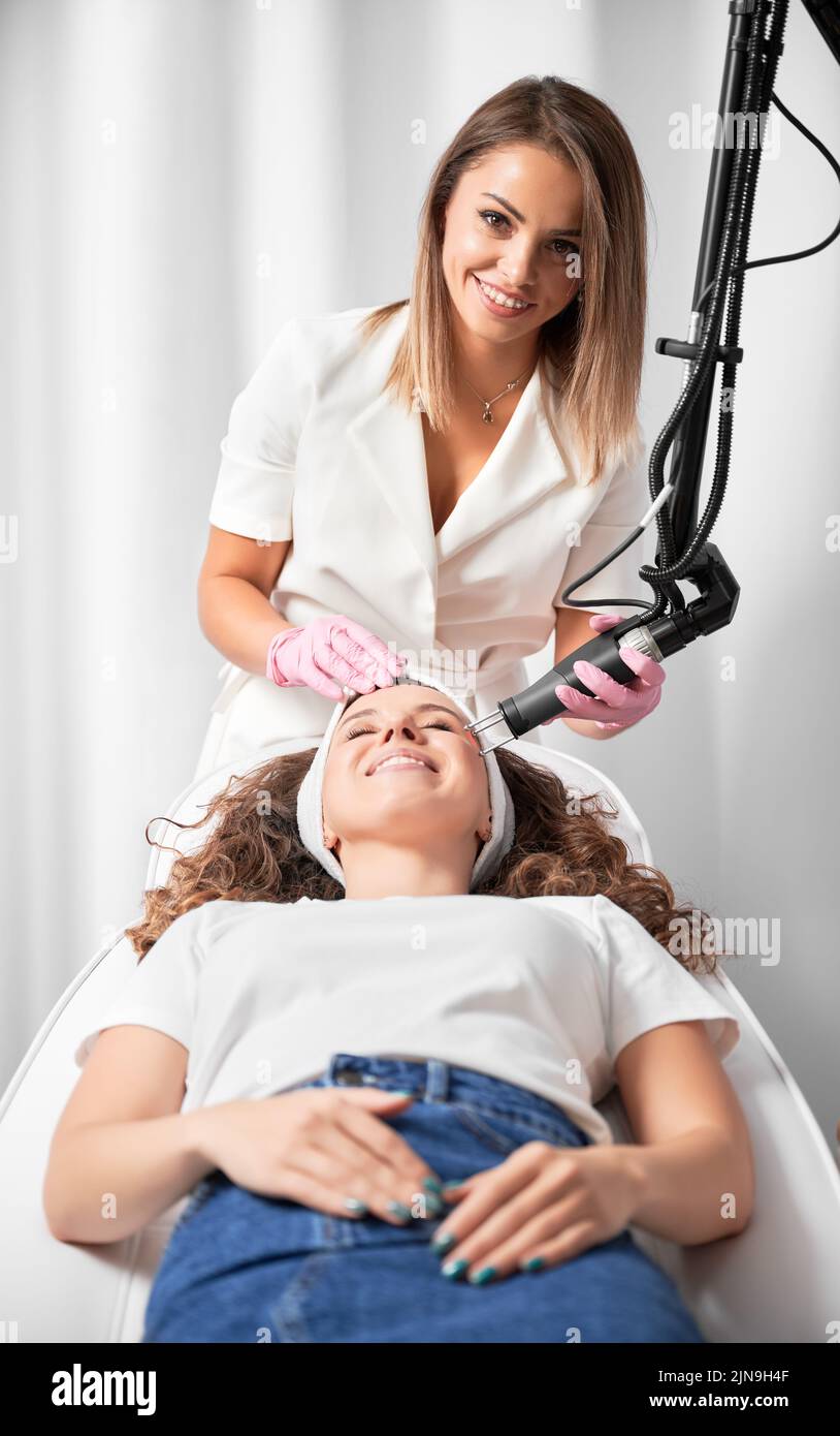 Portrait of smiling female cosmetologist performing laser skin resurfacing. Happy client in anticipation of skin rejuvenation after important procedure. Stock Photo