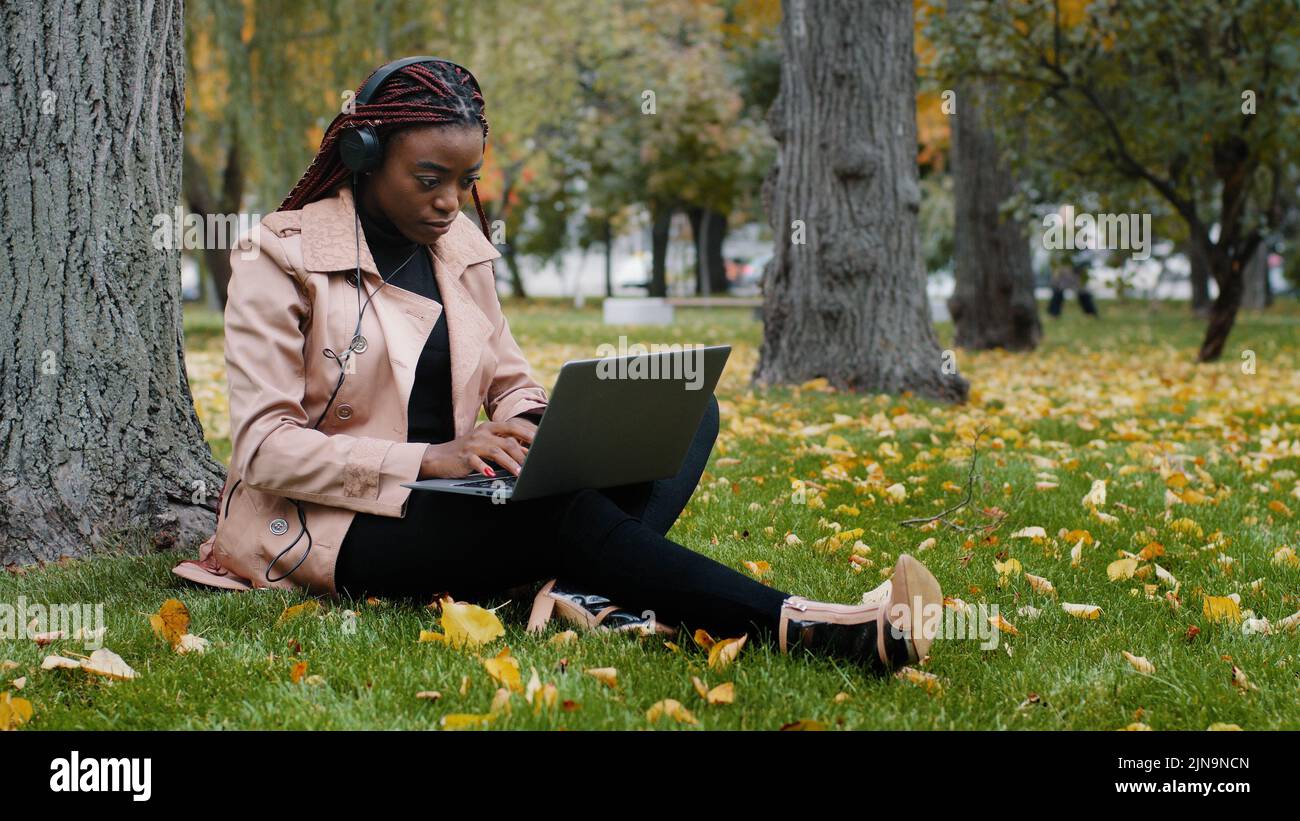 Young focused female student freelancer sitting in autumn park near tree working on laptop typing message checks email studying remote online distance Stock Photo