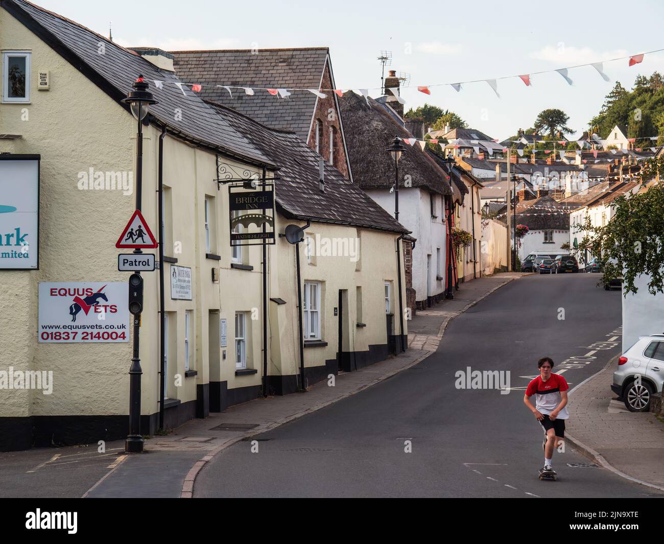 HATHERLEIGH, DEVON, ENGLAND - AUGUST 9 2022: View along Bridge St with cottages, bunting flags, and a young man on a skateboard, Stock Photo