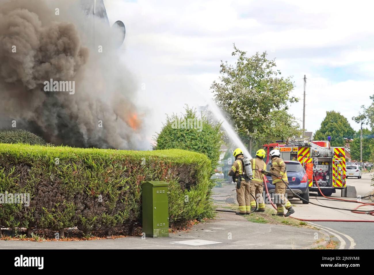Essex Fire and Rescue Service fire engine & group of fire brigade firemen firefighters spraying water jet on house fire smoke and flames   England UK Stock Photo