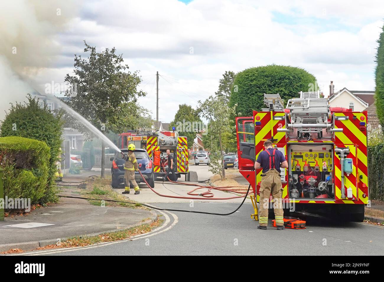 Firemen spraying water jet on house two fire brigade engines & firefighters Essex Fire and Rescue Service in residential street scene England UK Stock Photo