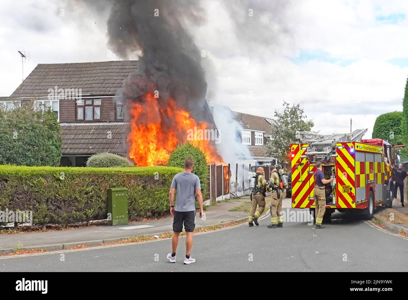 Flames & black smoke greet Essex Fire and Rescue services firemen arriving at house fire brigade fire engine & firefighters with spectator watching UK Stock Photo