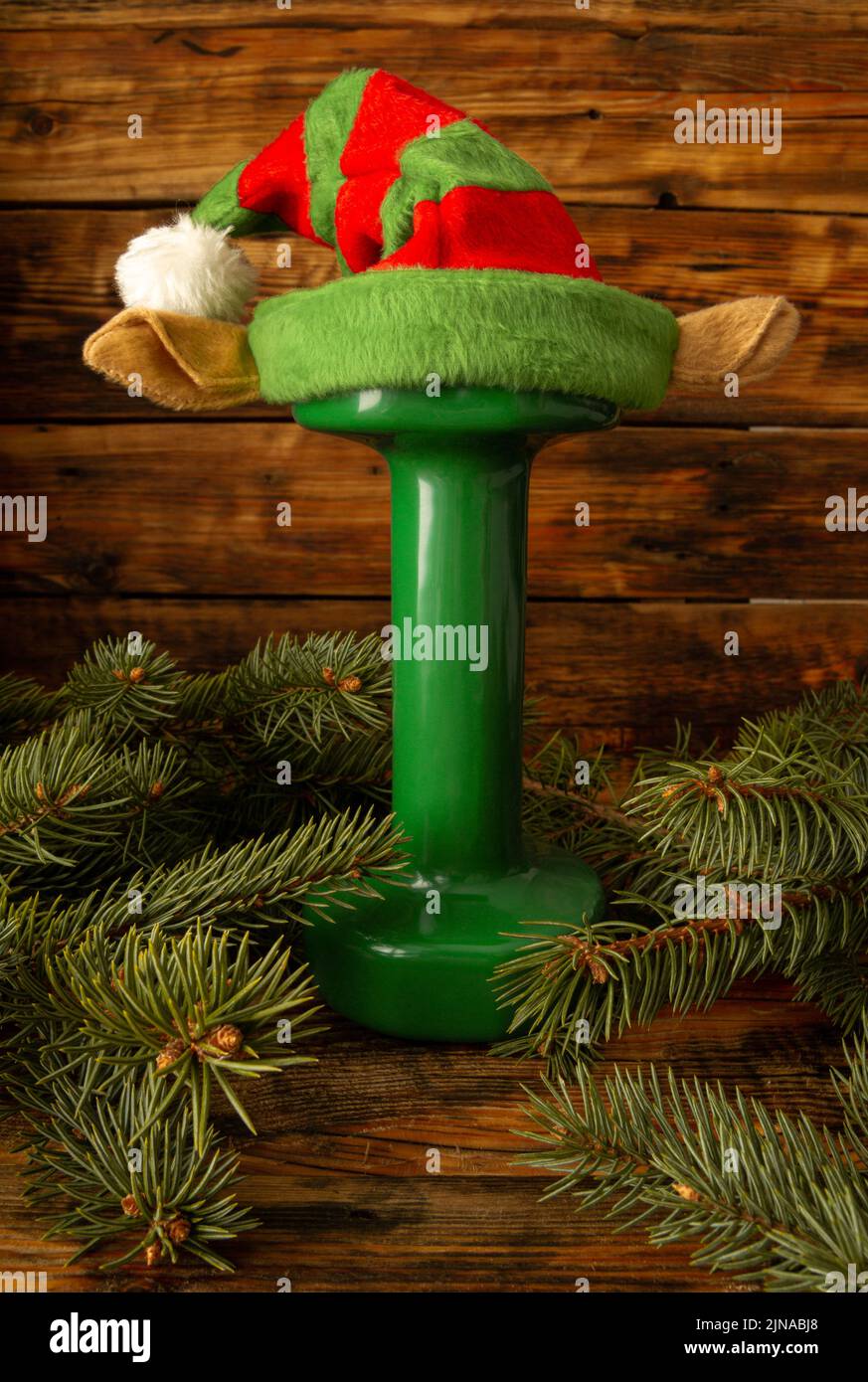 Green dumbbell with elf hat. Exercise equipment as a Christmas gift idea. Gym fitness holiday season concept, composition with tree branches. Stock Photo
