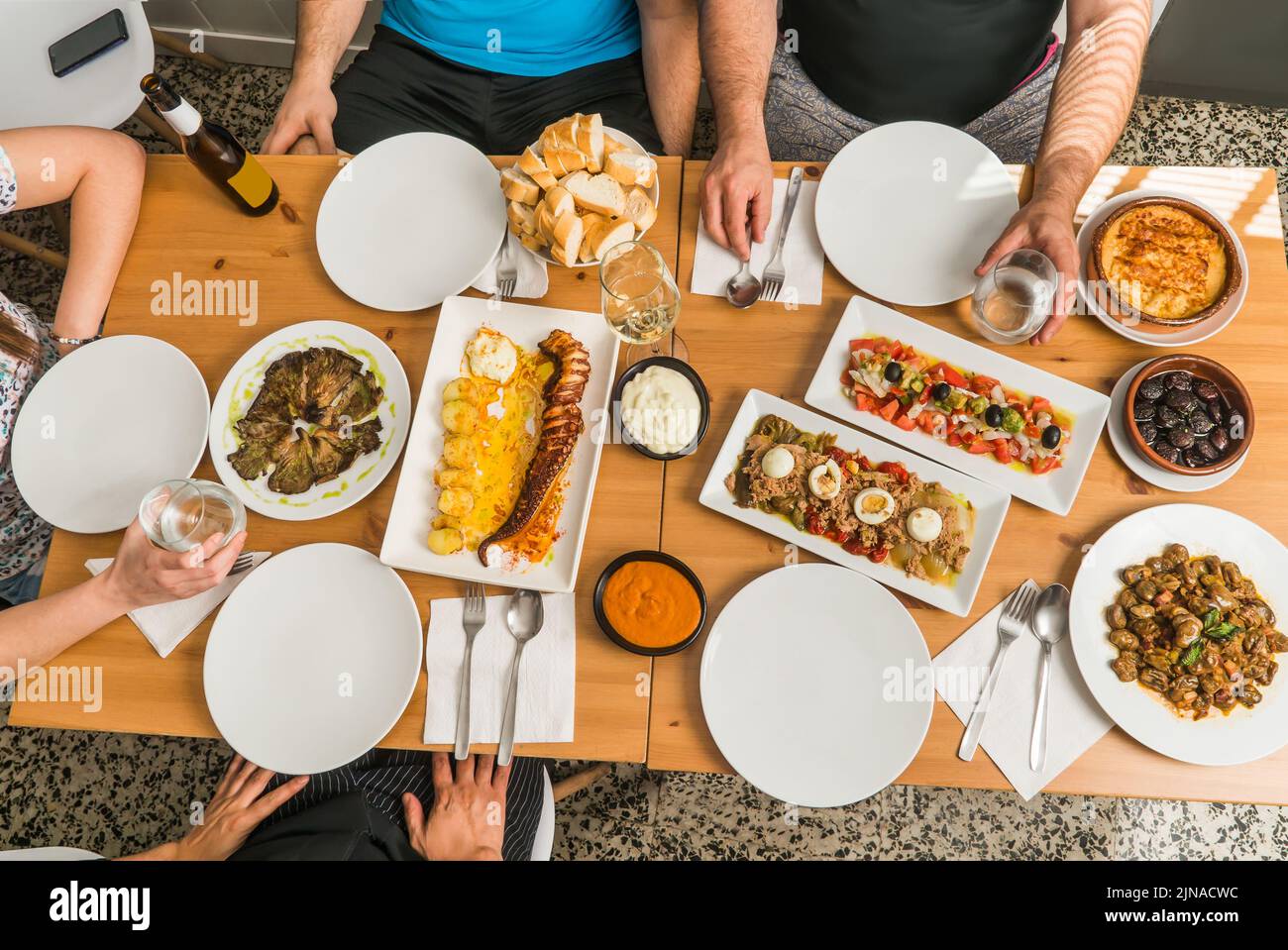 Table with tapas, salads and typical Spanish food around which people are sitting ready to eat. Focus on the center plates. Stock Photo