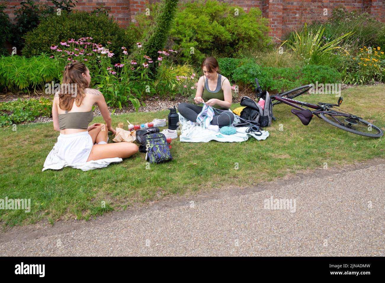 Young woman sitting on ground crocheting talking with friend by bike having picnic in summer 2022 Bute Park Cardiff Wales UK   KATHY DEWITT Stock Photo