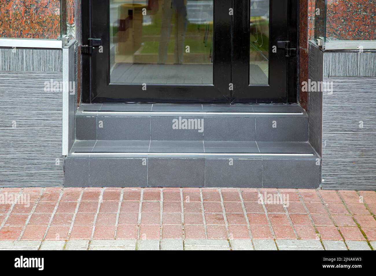pedestrian sidewalk near threshold from gray ceramic tile with steps at entrance to store with black door with glass, facade clad with tiles and grani Stock Photo