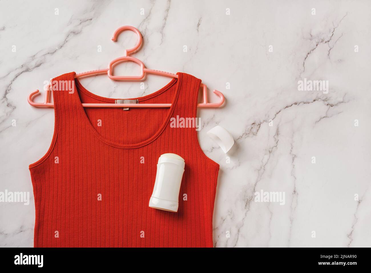 Red tank top on a hanger and solid antiperspirant over marble background. Deodorant stick on a linen jersey tank shirt. Prevent body odour, body care. Stock Photo