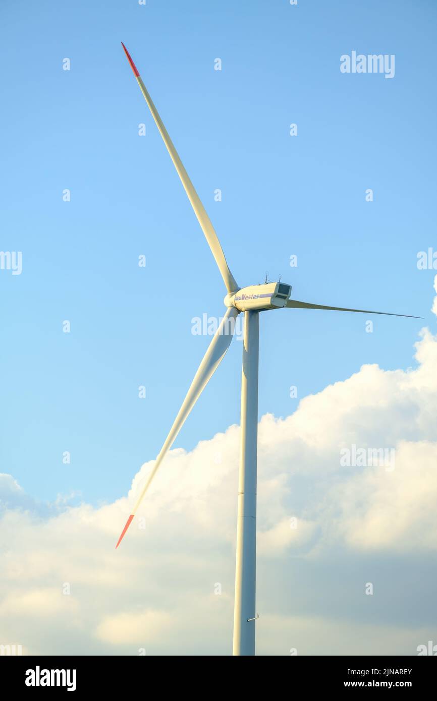 Windmill converts kinetic energy into electrical green energy against blue sky with white clouds. Long blades rotate producing energy closeup Stock Photo