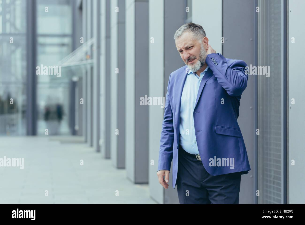 Injured and overtired senior gray-haired businessman, man outside office has severe neck pain, director holding hand massaging neck Stock Photo