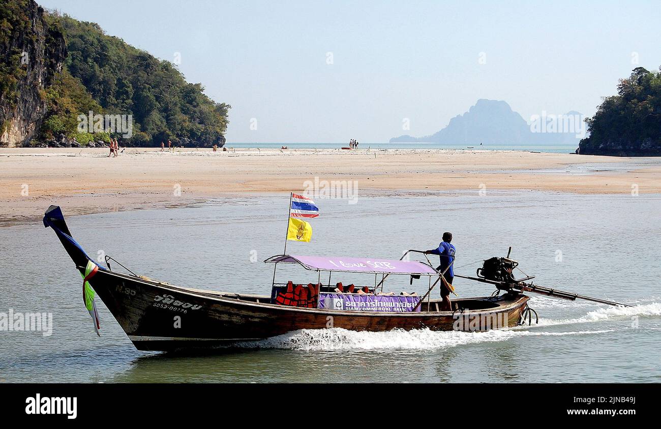 A man steers a longtail boat near Krabi in the Andaman Sea, Thailand, Asia. In the background is a beach on a mainland island with trees. Longtail boats are commonly used in Thailand as they often use secondhand car engines which are cheap to buy. Stock Photo