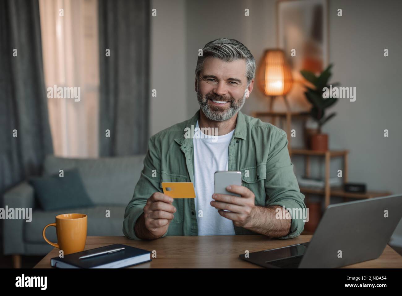 Buy and pay at home. Smiling retired european man with beard uses smartphone, computer and credit card Stock Photo