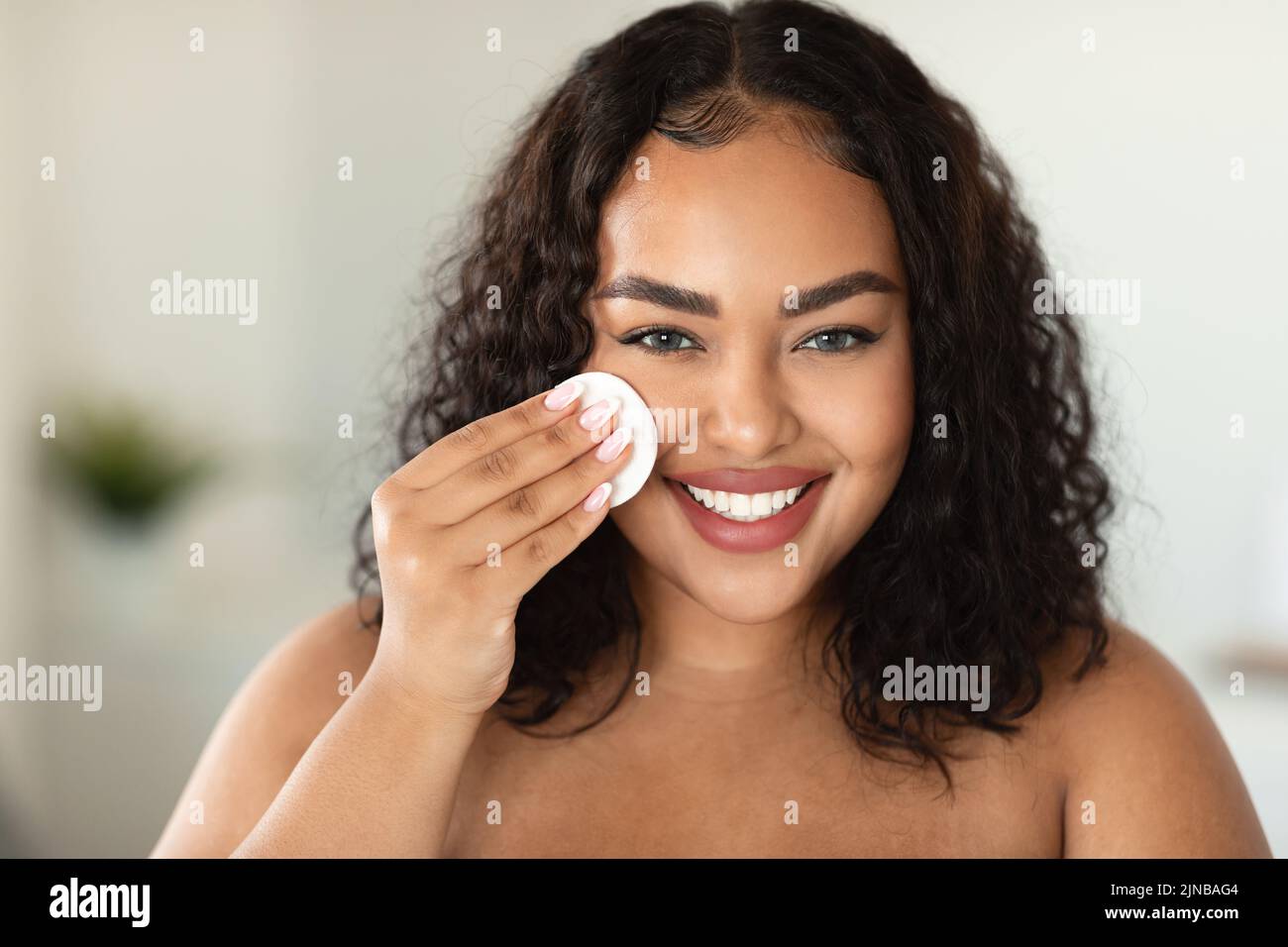 Makeup remover, cleansing and routine procedures. Happy african american oversize woman wiping face with cotton sponge Stock Photo