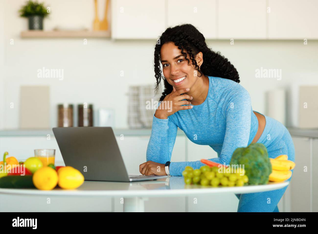 Searching for new healthy recipes. Happy fit lady cooking fresh salad at home in modern kitchen, using laptop Stock Photo