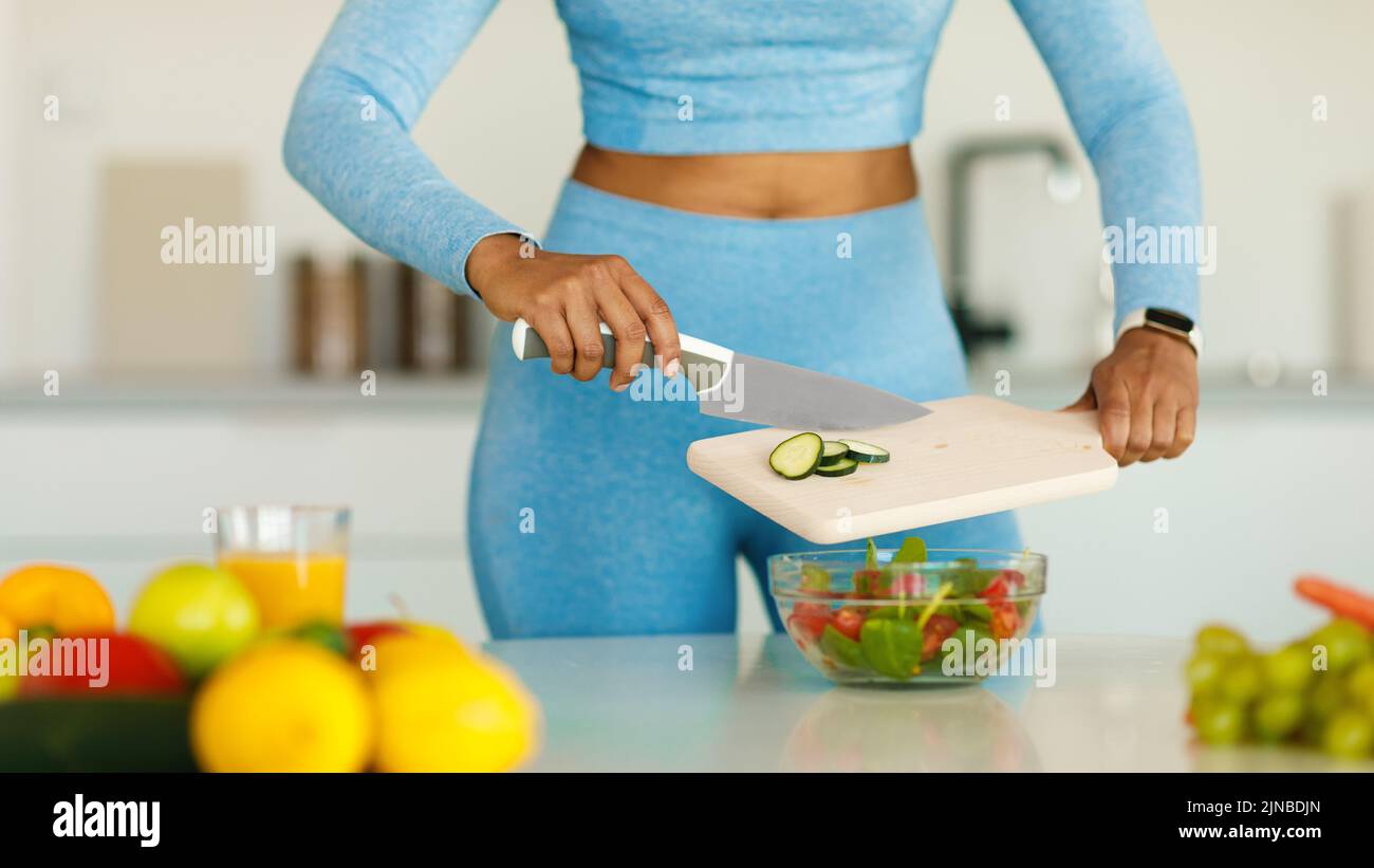 Detox recipes. Fit black lady cooking healthy vegetable salad, cutting cucumber on wooden board, cropped Stock Photo