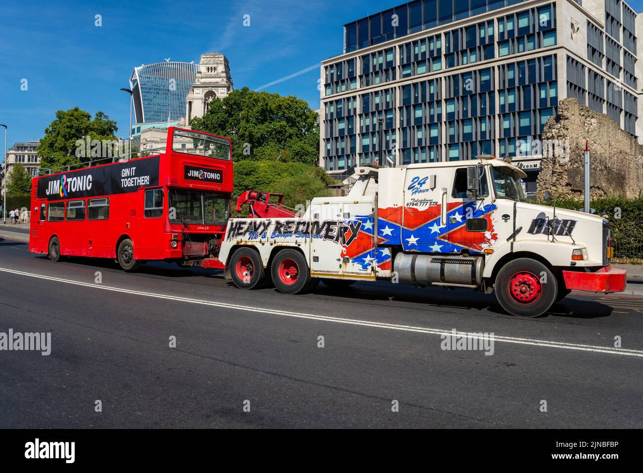 Heavy Recovery breakdown truck hauling a broken down bus. Jim and Tonic gin mobile bar bus being towed behind large American wrecking vehicle Stock Photo