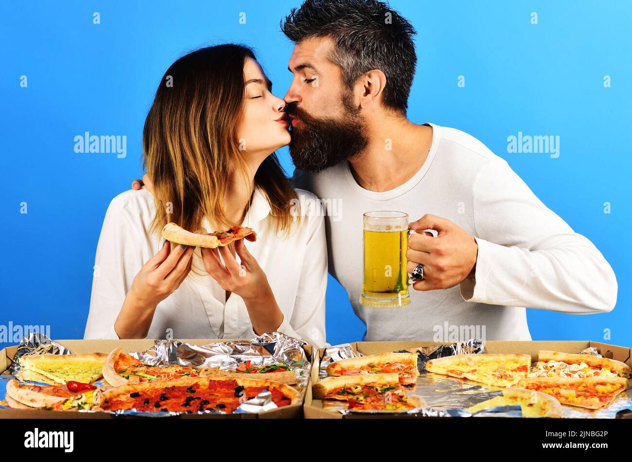 Couple sitting at table kissing and eating pizza. Woman with slice pizza and man with glass of beer. Stock Photo