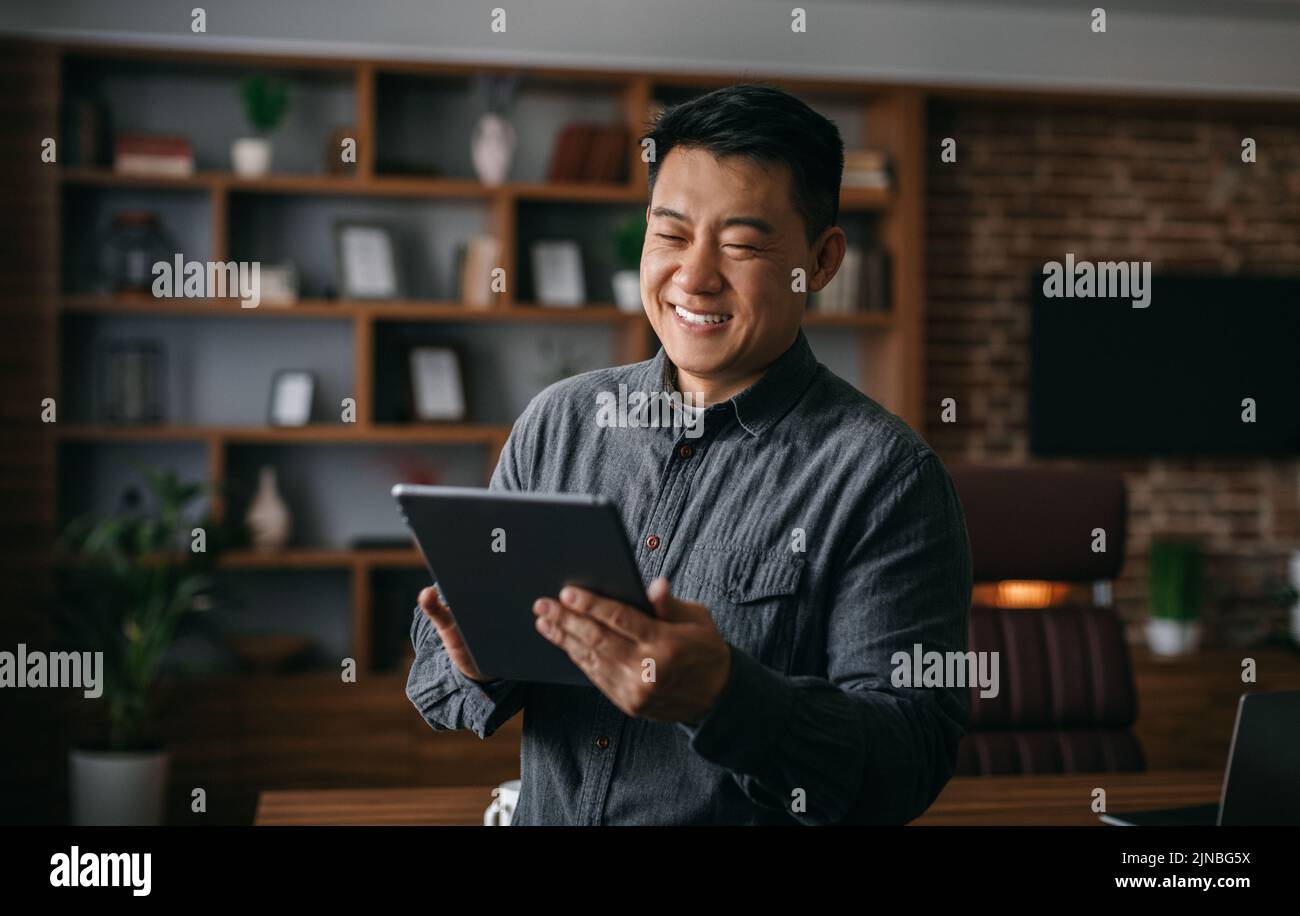 Smiling successful mature korean man typing on tablet, chatting with client, analyze startup data in office interior Stock Photo
