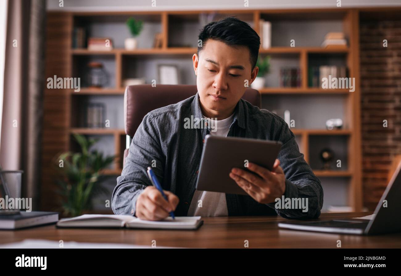 Busy concentrated mature chinese man at table with laptop uses tablet