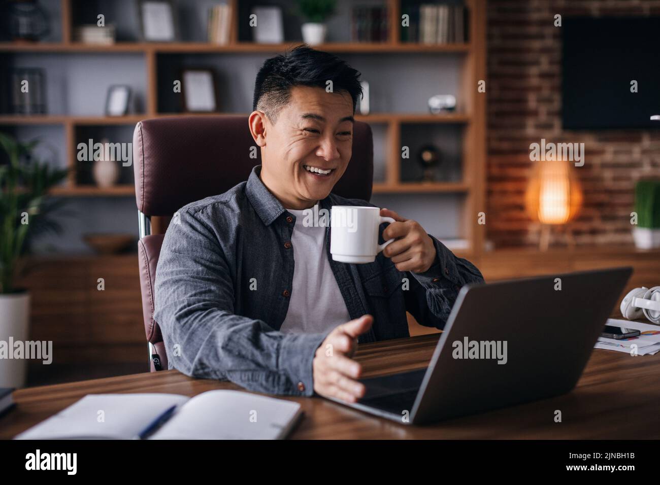 Glad adult chinese man ceo manager work with laptop with cup of drink and talking with client Stock Photo