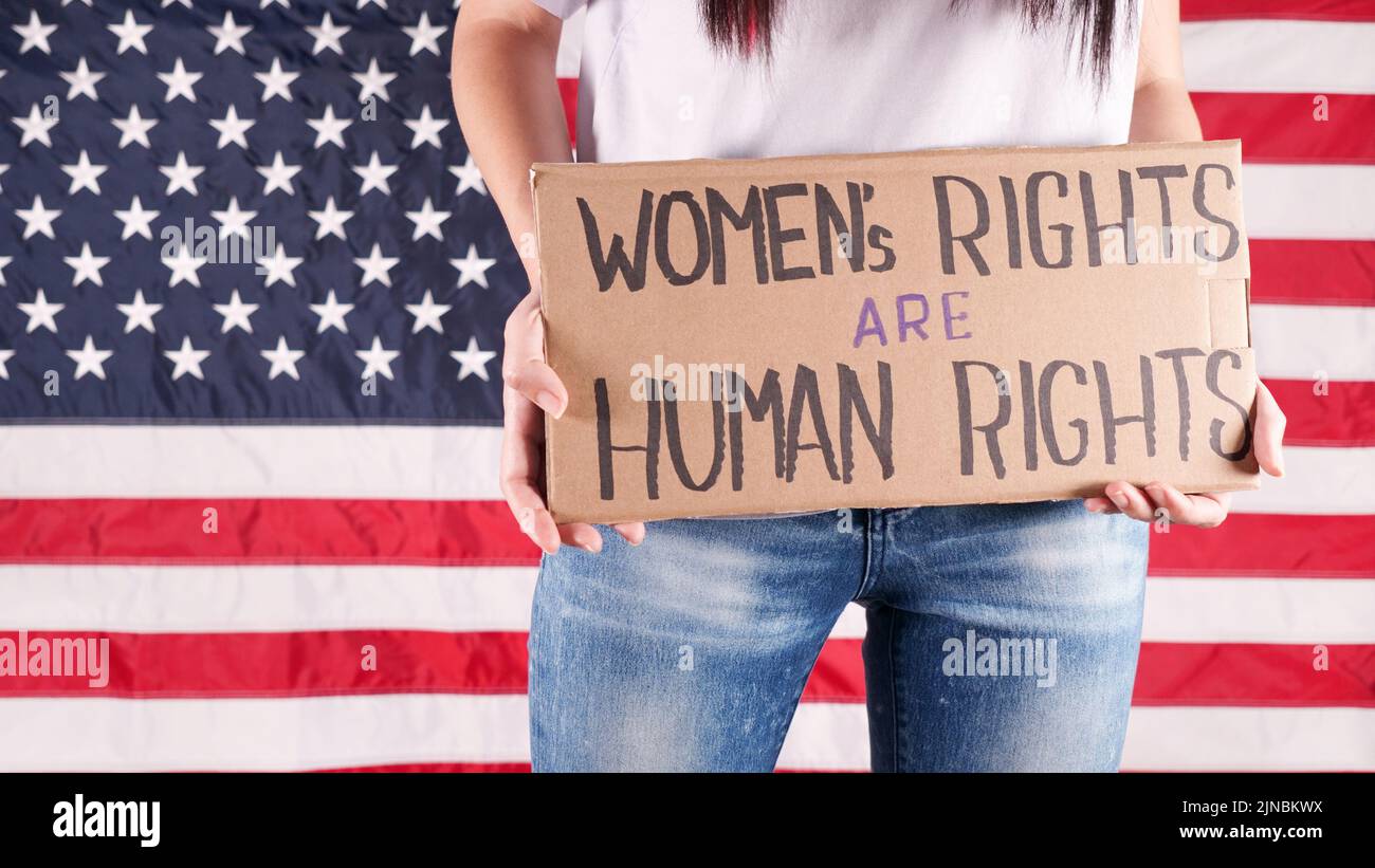 Young woman protester holds cardboard with Womens Rights Are Human Rights sign against USA flag on background. Feminist power. Equal opportunity Stock Photo