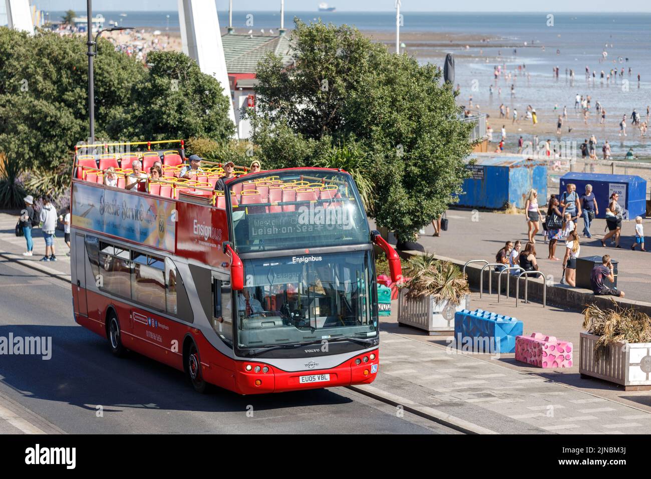 Open top red bus at the seaside on a sightseeing tour of Southend, with passenger on the top deck and tourists on the beach and promenade on a hot day Stock Photo