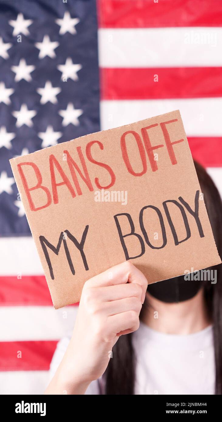 Young woman holding sign Bans Of My Body covering face stop abortion American flag on background. Anti domestic violence, racism racial discrimination Stock Photo