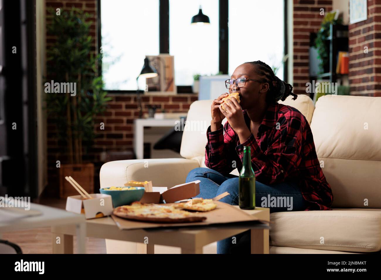 African american woman eating burger from fast food delivery takeout, watching movie on tv channel program. Serving takeaway meal to enjoy leisure activity with film on television. Stock Photo