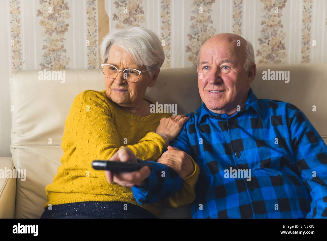 Senior modern couple actively watching a scary film or news on a tv, a man holding a remote controler. High quality photo Stock Photo